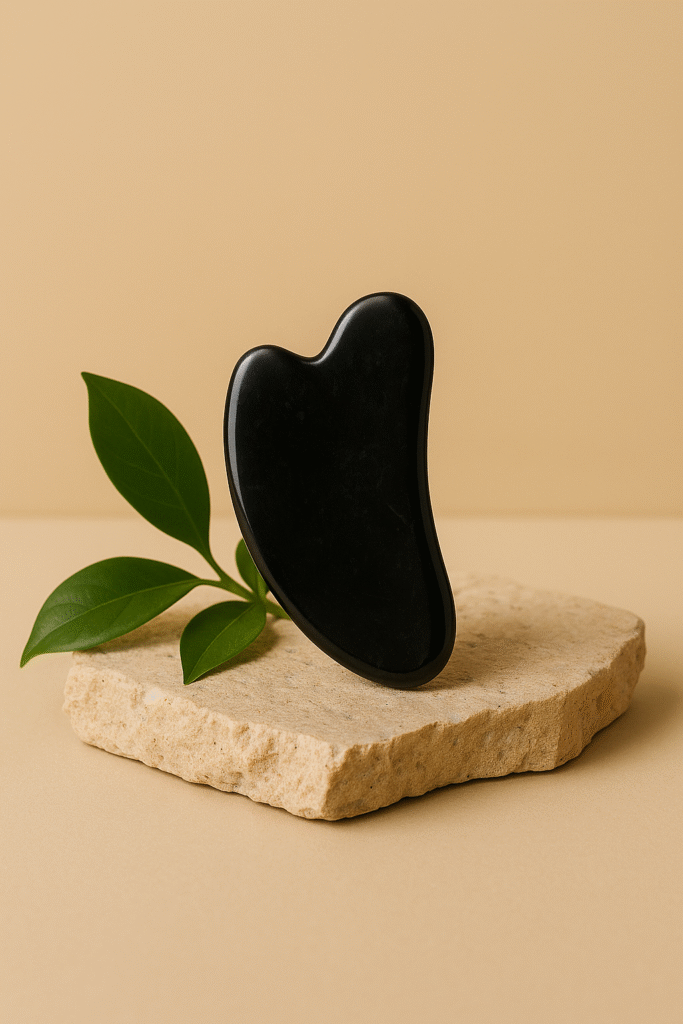 Black obsidian gua sha stone standing on a textured stone slab with green leaves on a beige background.